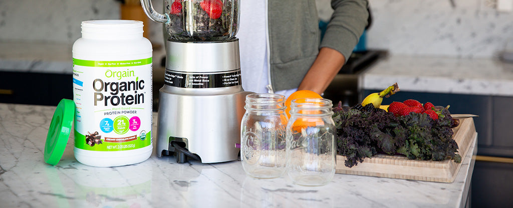 A person prepares a smoothie in a kitchen. On the marble counter are a blender with strawberries, a jar of Orgain Organic Protein powder, two empty mason jars, and a tray of fresh fruits and vegetables, including bananas, kale, oranges, and strawberries.