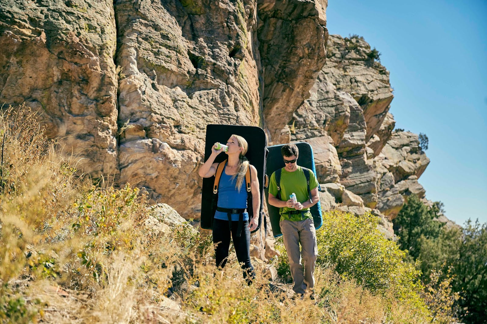 Two rock climbers take a break, holding drinks, with climbing mats on their backs and rocky hillside in the background.