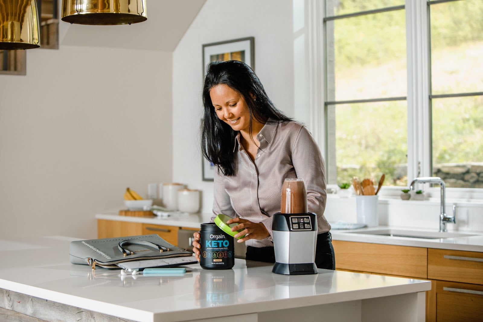 A woman with dark hair prepares a smoothie in a modern kitchen, smiling and holding a green-lidded "Keto" container.