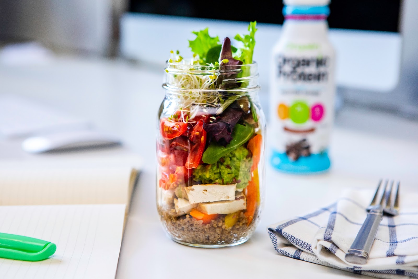 A fresh salad in a glass jar, layered with quinoa, tofu, colorful vegetables, and leafy greens, is placed on a white desk next to an open notebook with a green pen. A bottle of Orgain Organic Protein shake and a plaid napkin with a fork are also visible.