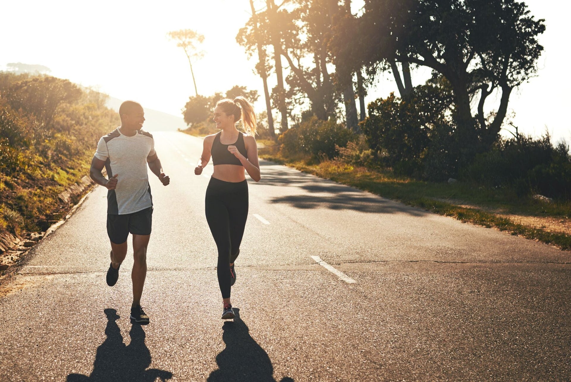 A man and woman jogging on a sunlit road lined with trees and greenery, conversing and enjoying the run.