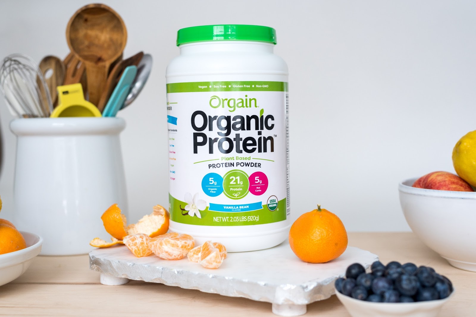 A container of Orgain Organic Protein powder (Vanilla Bean flavor) on a countertop with peeled and whole oranges, a bowl of blueberries, and a utensil holder with kitchen utensils.