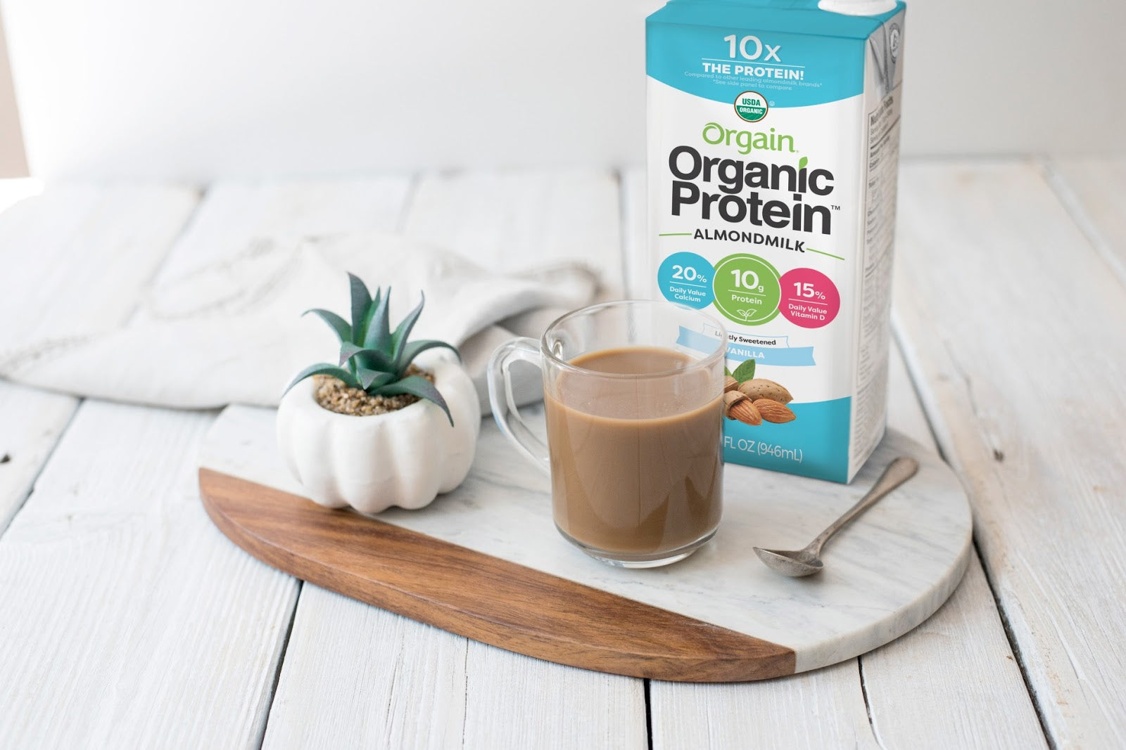 A carton of Orgain Organic Protein Almond Milk is placed on a marble and wooden cutting board next to a clear glass mug filled with the milk. A small potted succulent and a spoon are also on the board, set on a light wooden surface.