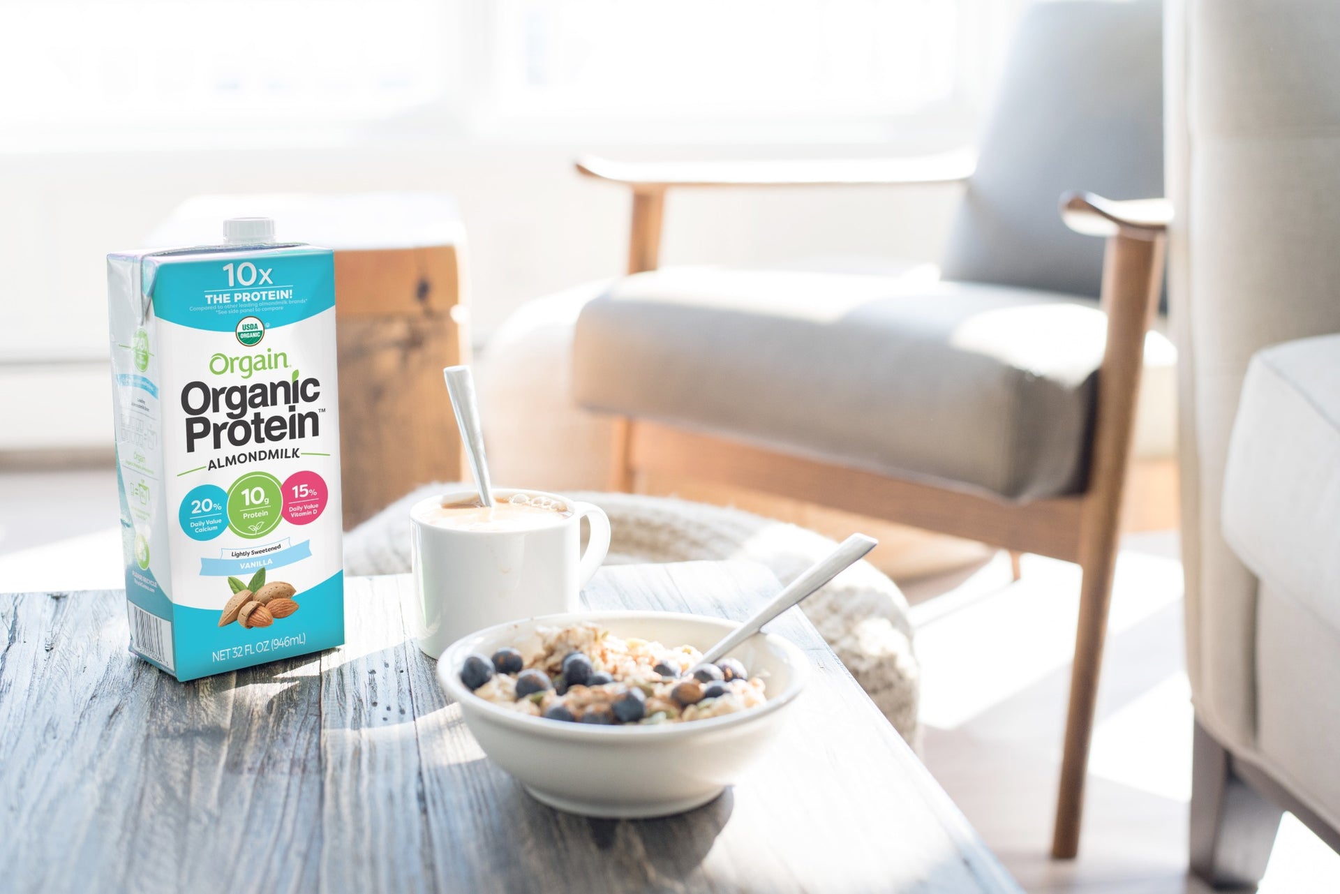 A carton of Orgain Organic Protein Almond Milk sits on a wooden coffee table beside a bowl of granola and berries and a cup of coffee with a spoon. In the background, a wooden chair with gray cushions and a white sofa are visible in a brightly lit room.