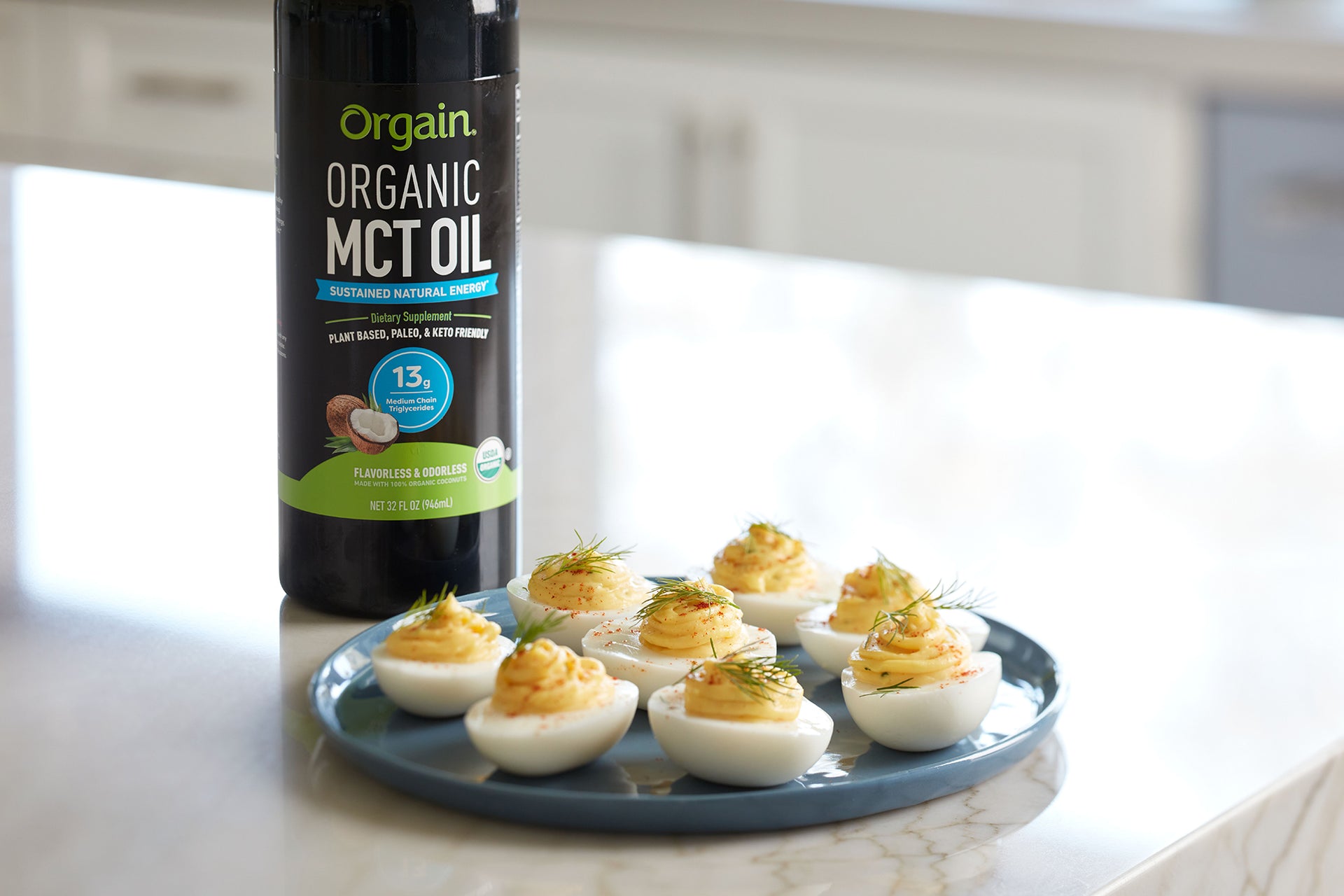 A bottle of Orgain Organic MCT Oil is placed on a white counter beside a blue plate filled with eight deviled eggs. The deviled eggs are garnished with herbs, and the overall scene is bright and light-filled, indicating a kitchen setting.