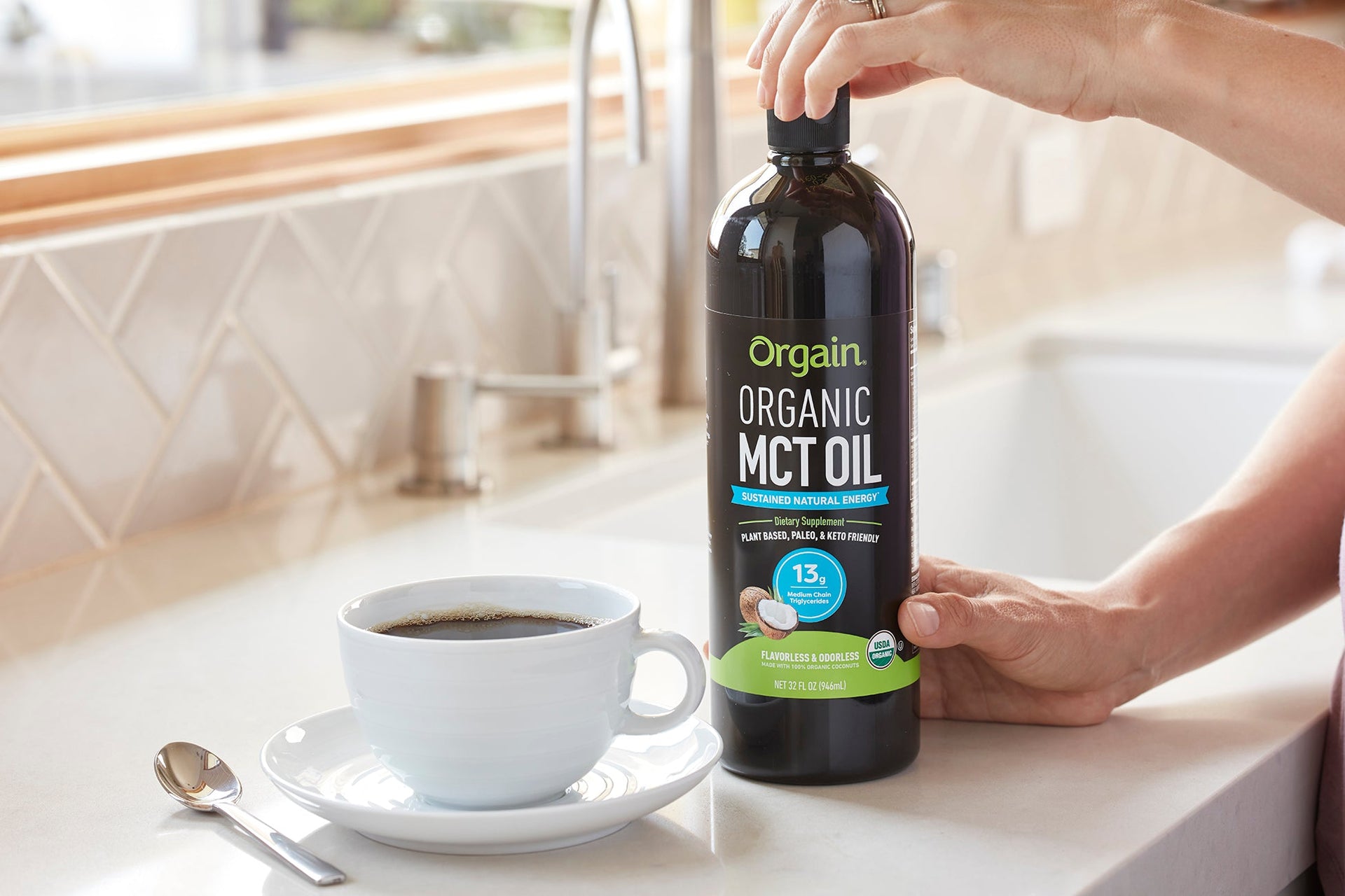 A hand holds a bottle of Orgain organic MCT oil next to a cup of coffee on a kitchen countertop.