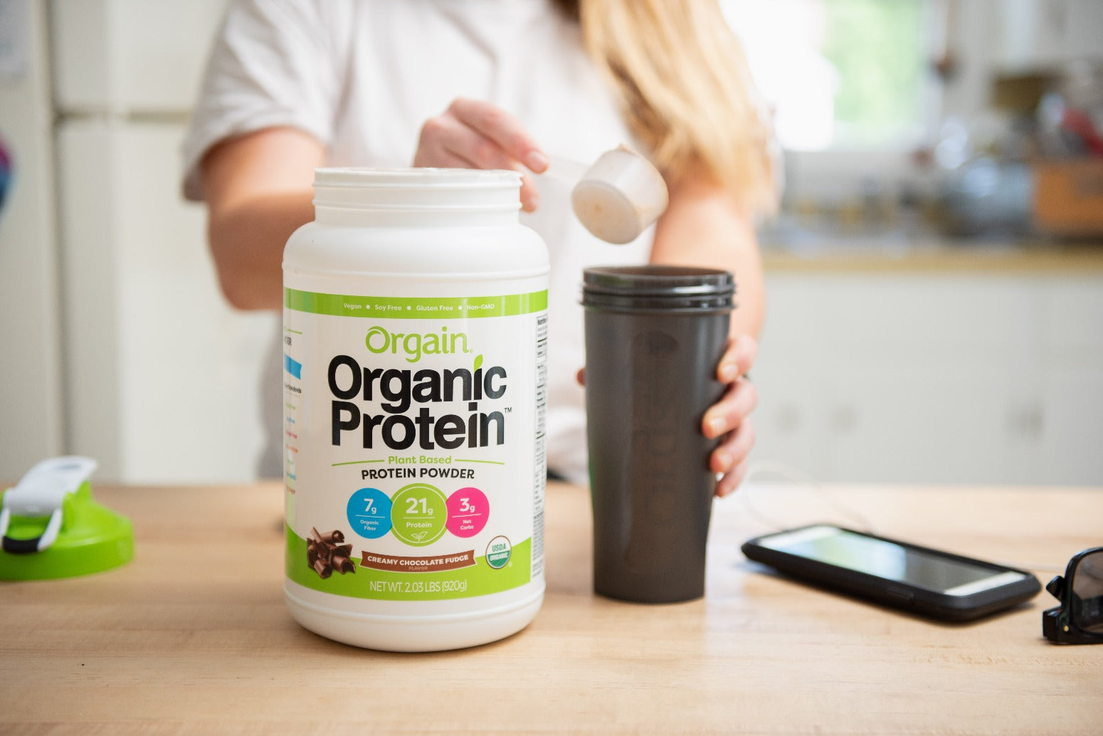Person scooping Orgain Organic Protein Powder into a black shaker bottle. The protein powder container is prominently displayed on a kitchen counter. Nearby items include a phone, sunglasses, and another lid or shaker component.