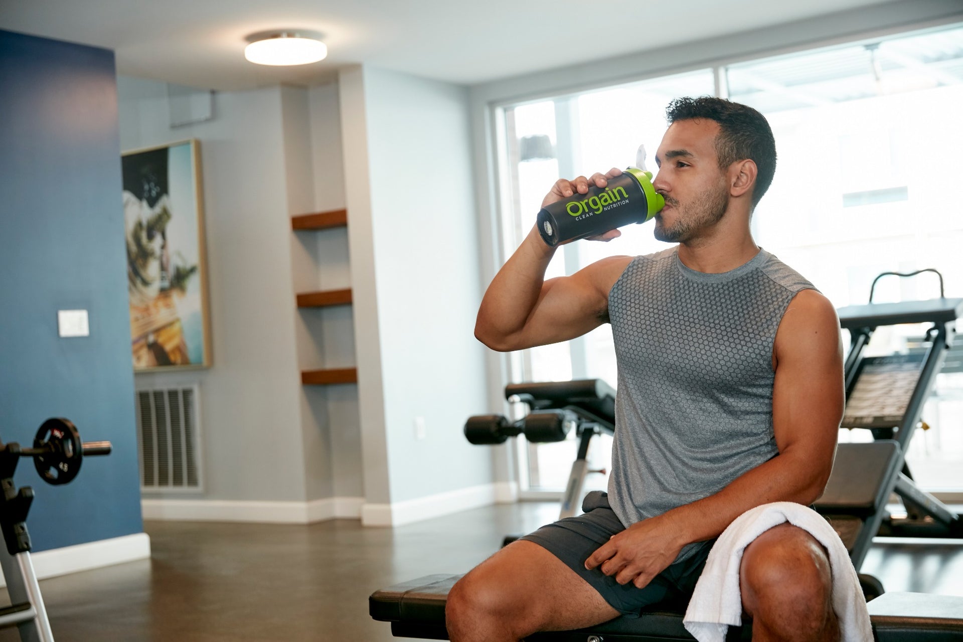 A person in athletic attire sits on a gym bench, drinking from a black water bottle labeled "Orgain."