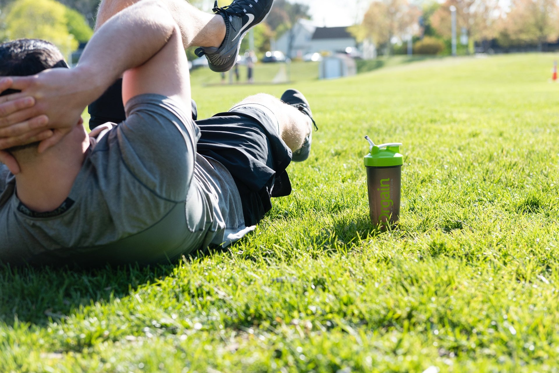 A person in athletic wear is lying on their back on a grassy field, performing abdominal exercises. There is a green and black water bottle placed upright on the grass near them. In the background, there are trees and buildings under a sunny sky.