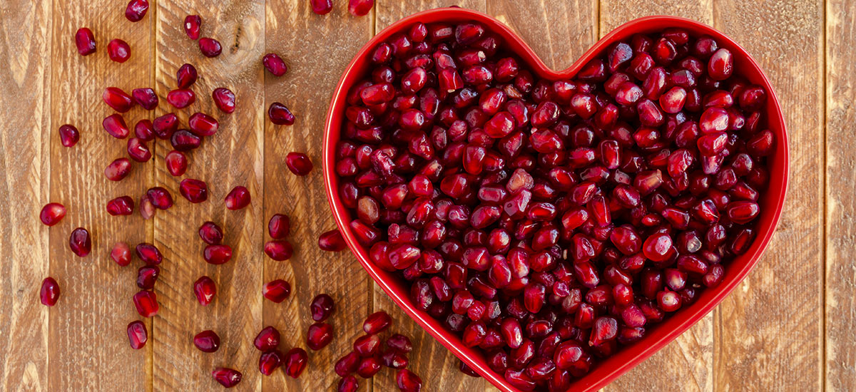 A heart-shaped bowl filled with bright red pomegranate seeds sits on a wooden surface. Several seeds are scattered around the bowl, adding a playful touch to the arrangement. The vibrant color of the seeds contrasts with the rustic wood background.