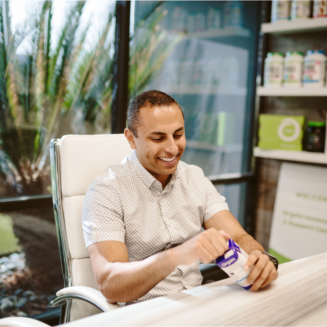 A person in a light-colored shirt smiles while opening a container of vitamins, with large windows and shelves in the background.
