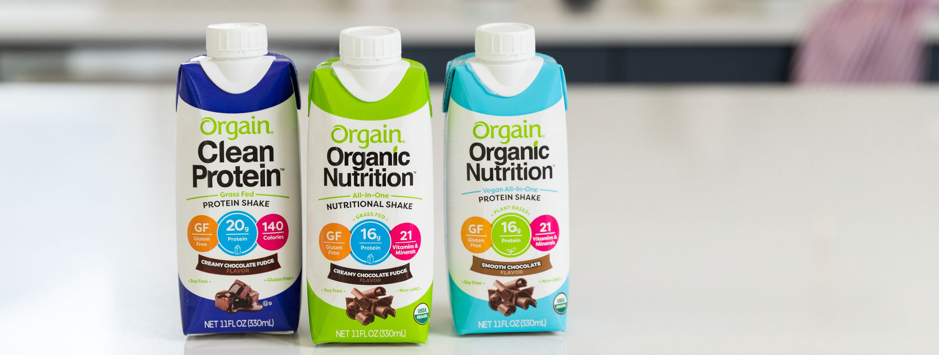 Three cartons of Orgain nutritional drinks, each labeled Creamy Chocolate Fudge, displayed side by side on a kitchen counter.