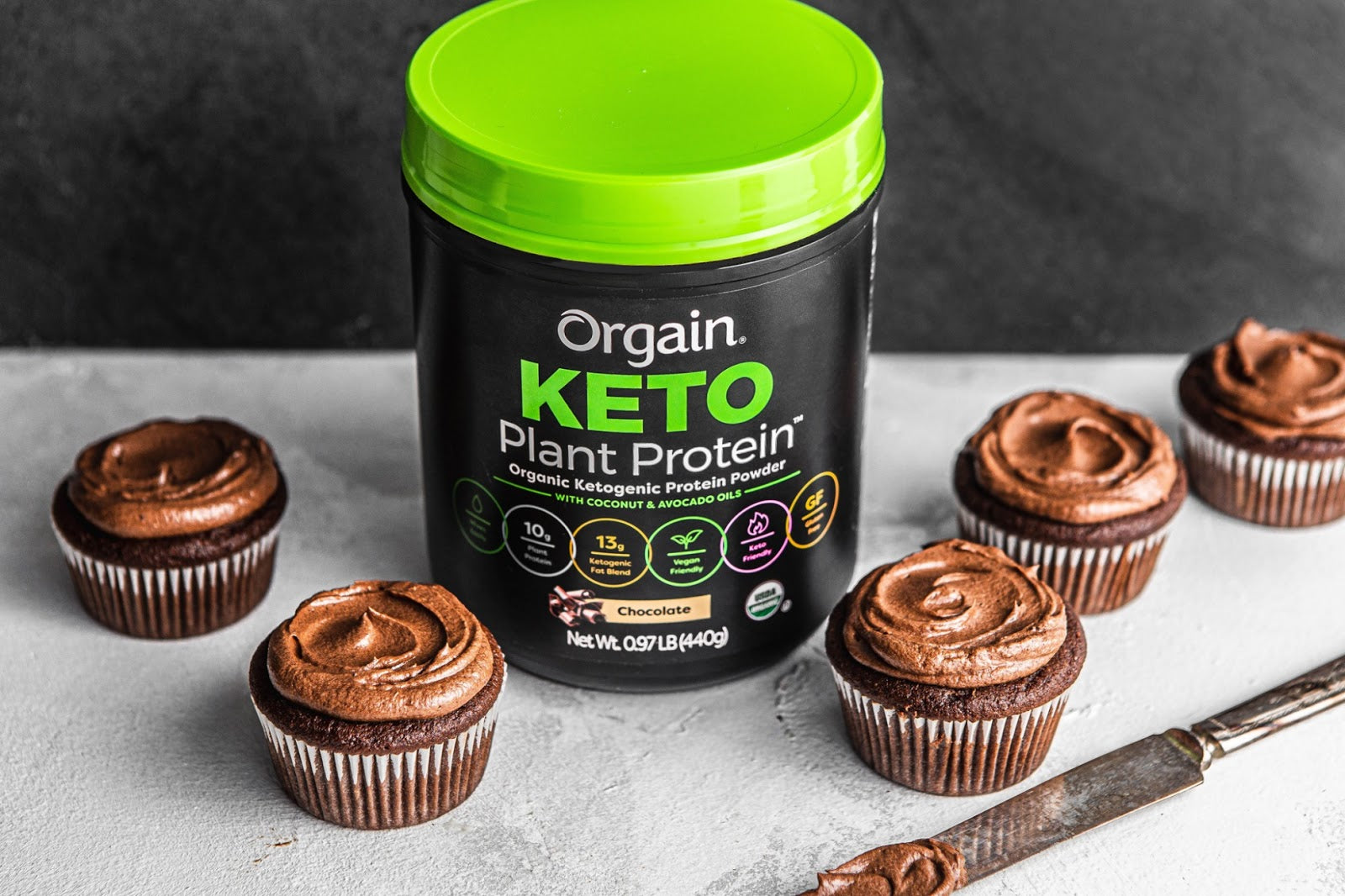Orgain Keto Plant Protein powder surrounded by chocolate cupcakes. A knife with frosting on the blade lies nearby.