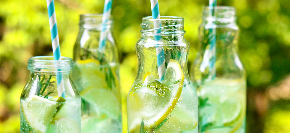 Four glass bottles filled with sparkling lemon water and garnished with lemon slices and fresh herbs. Each bottle has a blue-and-white striped straw. The vibrant, green, outdoor background suggests a refreshing summertime beverage setting.