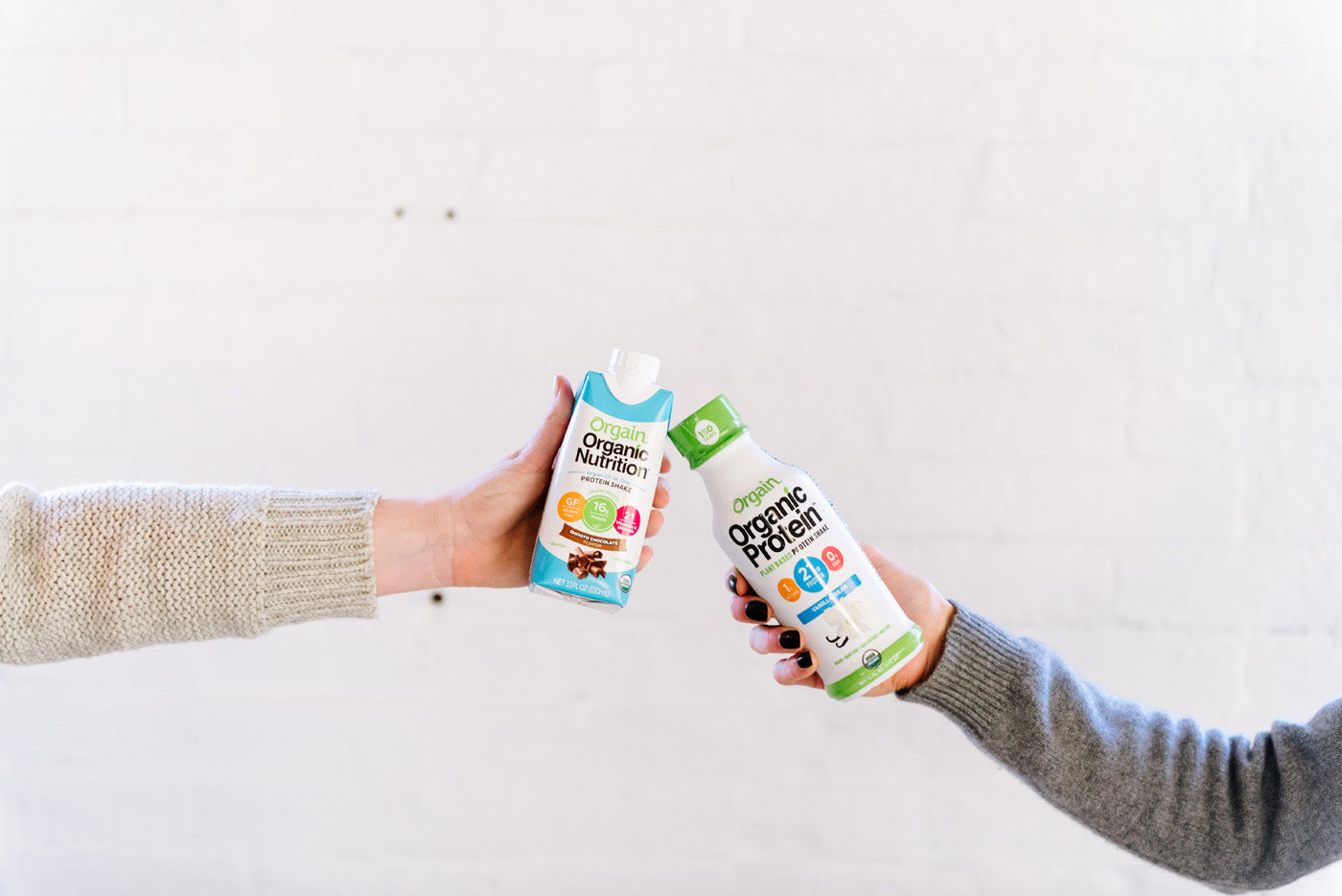 Two hands are pictured holding different Orgain nutritional beverages against a white brick wall. The left hand holds a carton of Orgain Organic Nutrition in chocolate flavor, and the right hand holds a bottle of Orgain Organic Protein in vanilla flavor.