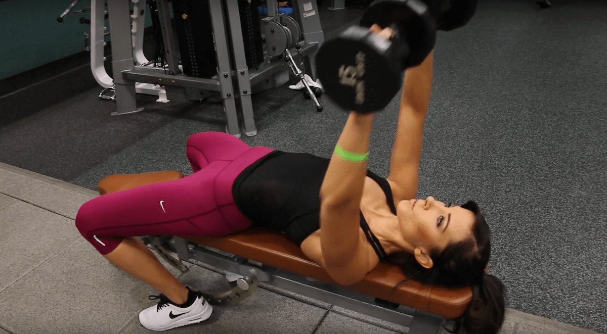 A woman in a black tank top and bright pink leggings lies on a bench press at the gym. She is lifting two dumbbells, performing a chest press exercise. Gym equipment and weights are visible in the background.