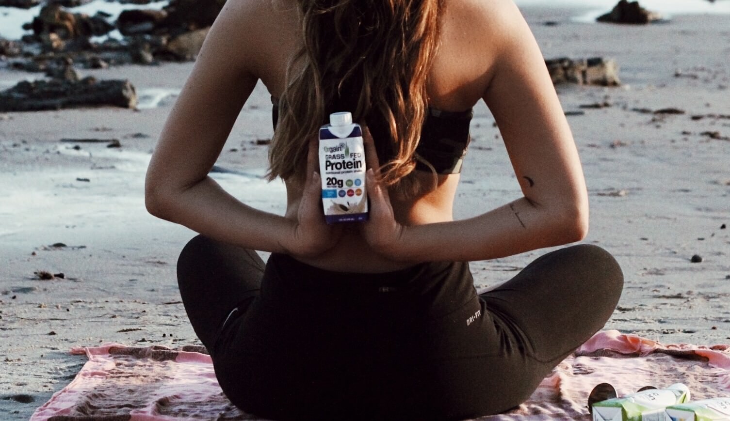 A person in a black sports bra and leggings sits cross-legged on a pink yoga mat at the beach, holding a shake bottle.