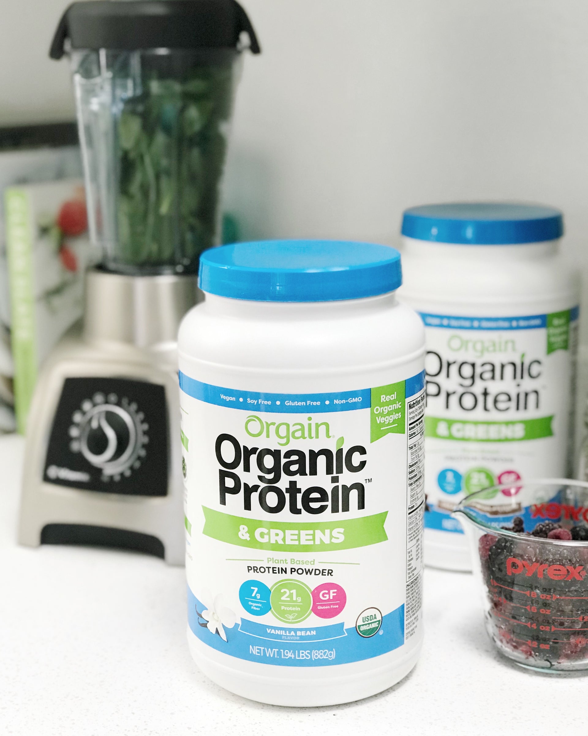 Two tubs of Orgain's Organic Protein powder on the kitchen counter next to a blender.