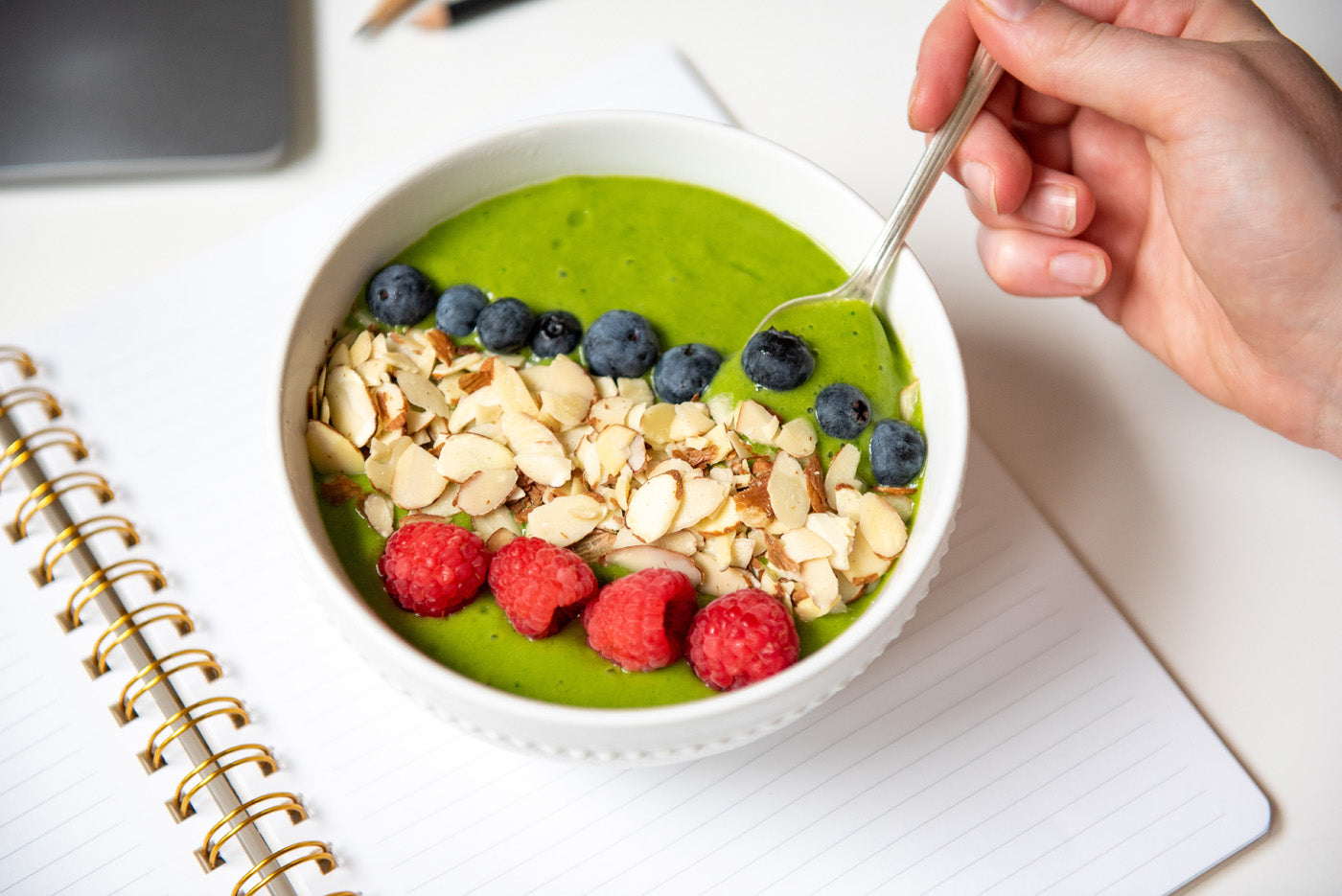 A hand holding a spoon over a bowl of green smoothie topped with sliced almonds, blueberries, and raspberries. The bowl is on top of an open spiral notebook with lined pages. A laptop and a pencil are visible in the background.