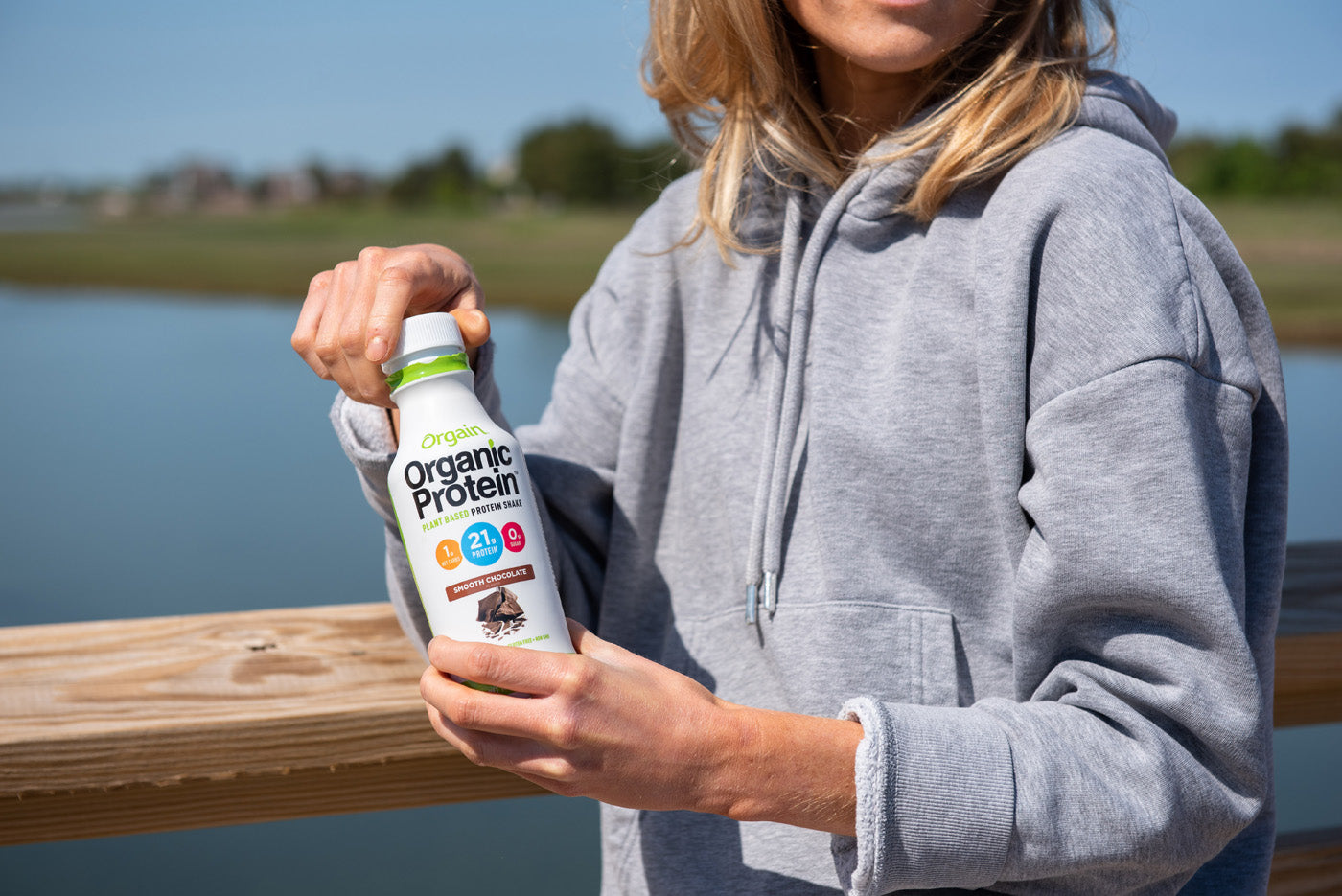 A person wearing a grey hoodie is holding a bottle of Orgain Organic Protein shake with both hands. They are standing outdoors on a wooden structure with a body of water in the background.