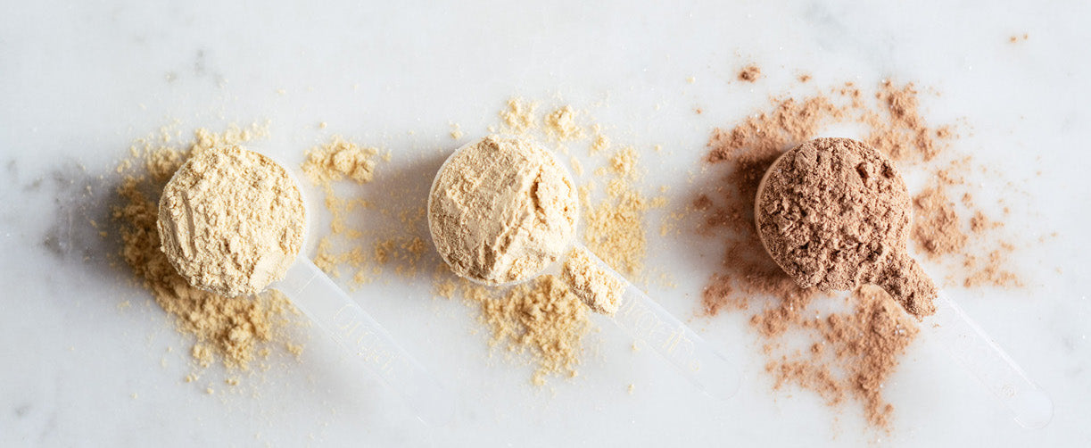 Three scoops of different protein powders, each with a bit of powder spilled around them, on a white marble surface. From left to right: one light yellow, one beige, and one brown. Each scoop has a transparent handle.