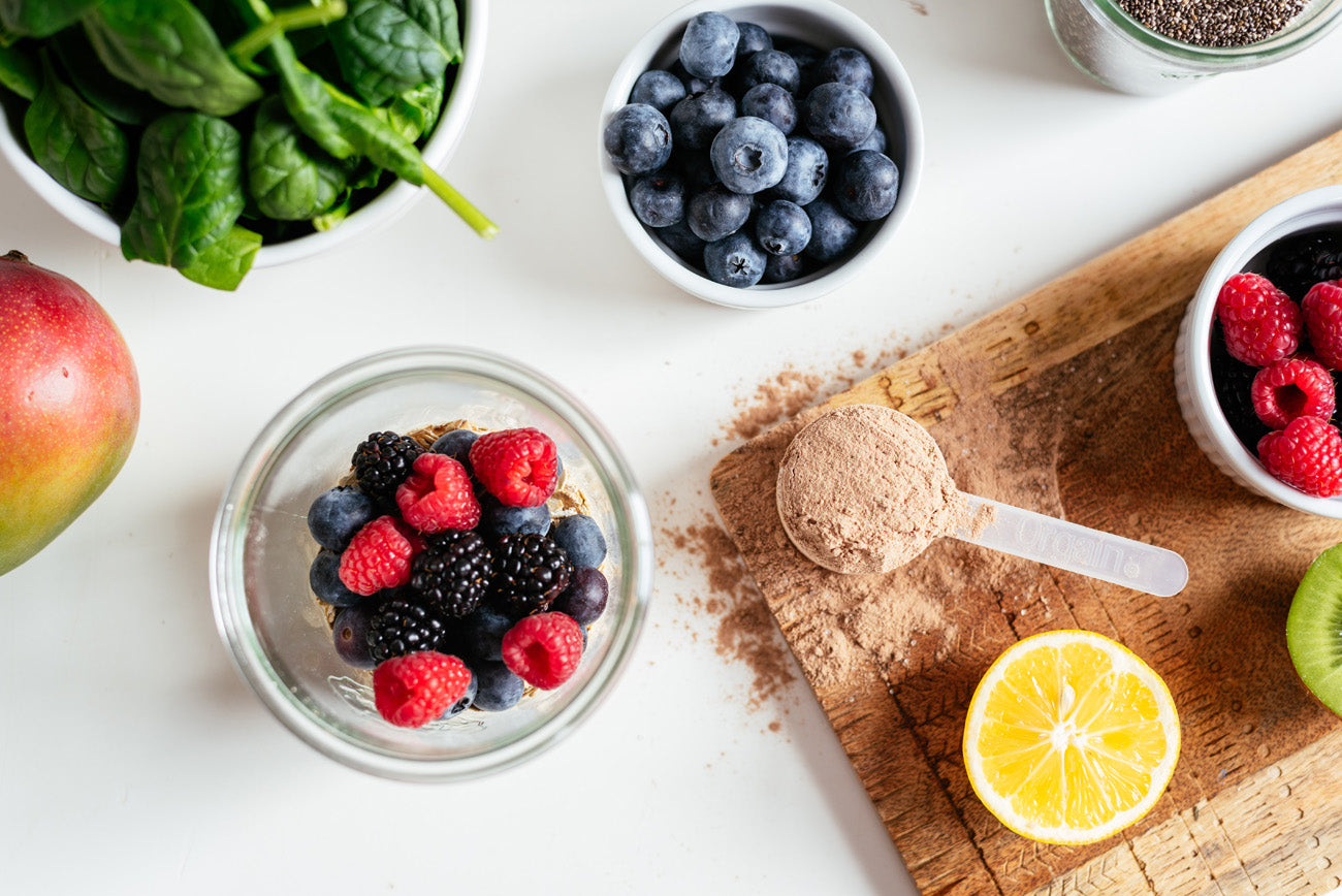 A healthy breakfast setup featuring a cutting board with chocolate protein powder, a half lemon, kiwi slices, and bowls of blueberries, blackberries, raspberries, and spinach leaves. A glass jar of layered berries and cereal is placed in the center.