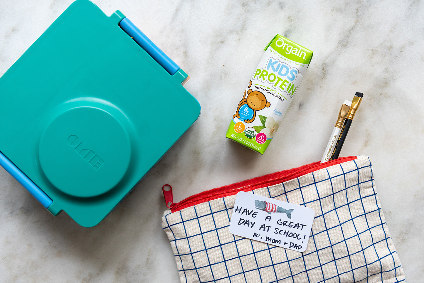 A teal lunchbox, an Orgain Kids Protein drink, a blue and red grid-patterned pouch with pencils, and a note saying, "Have a great day at school! XO, Mom & Dad" are arranged on a marble surface.