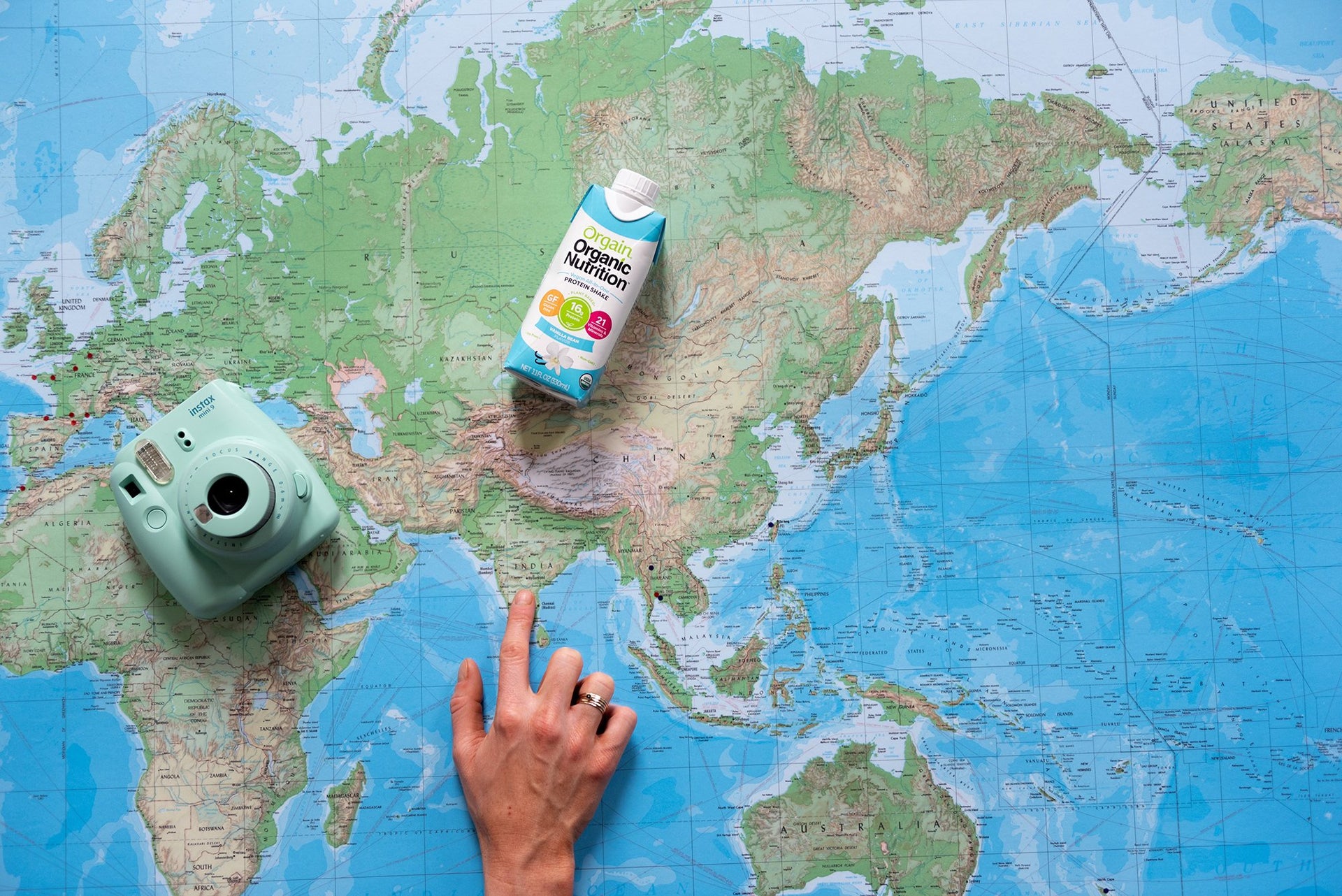 A hand pointing at a location on a world map, with a camera on the left side and a carton of organic coconut milk resting above the hand. The map shows continents of Europe, Asia, and part of Africa.
