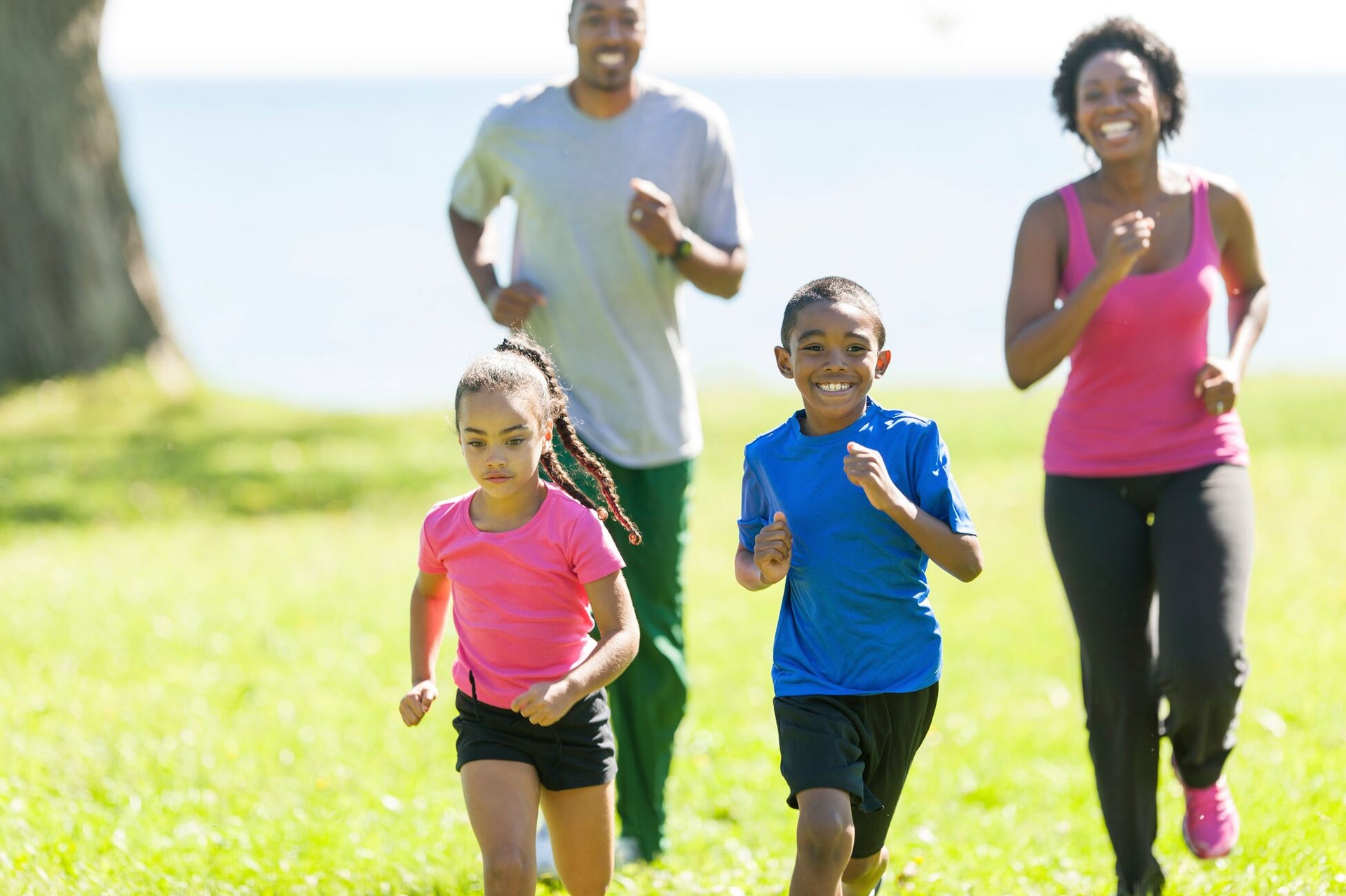 A joyous family of four, including two children and two adults, runs together on a grassy area near a body of water. Both children are in the foreground wearing colorful athletic clothes, while the adults follow closely behind, smiling.