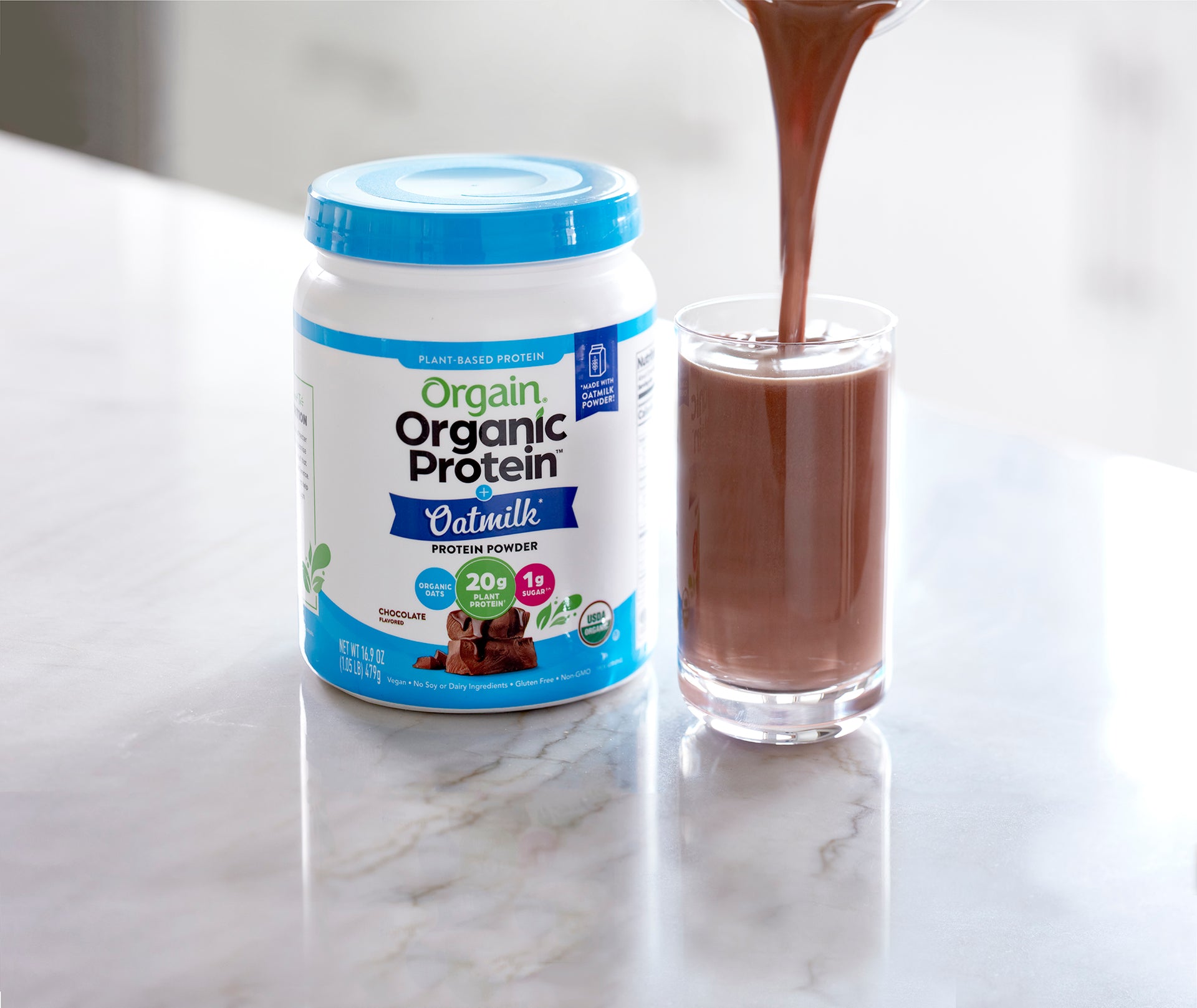 Orgain Organic Protein Oatmilk Powder on a kitchen counter next to a glass being filled with a protein shake.