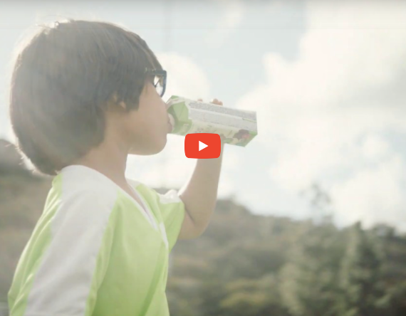 A child with short dark hair and glasses is drinking from a small carton outdoors. The child is wearing a green and white shirt. The background shows a blurred natural setting with trees and a cloudy sky. The YouTube play button is overlaid on the image.