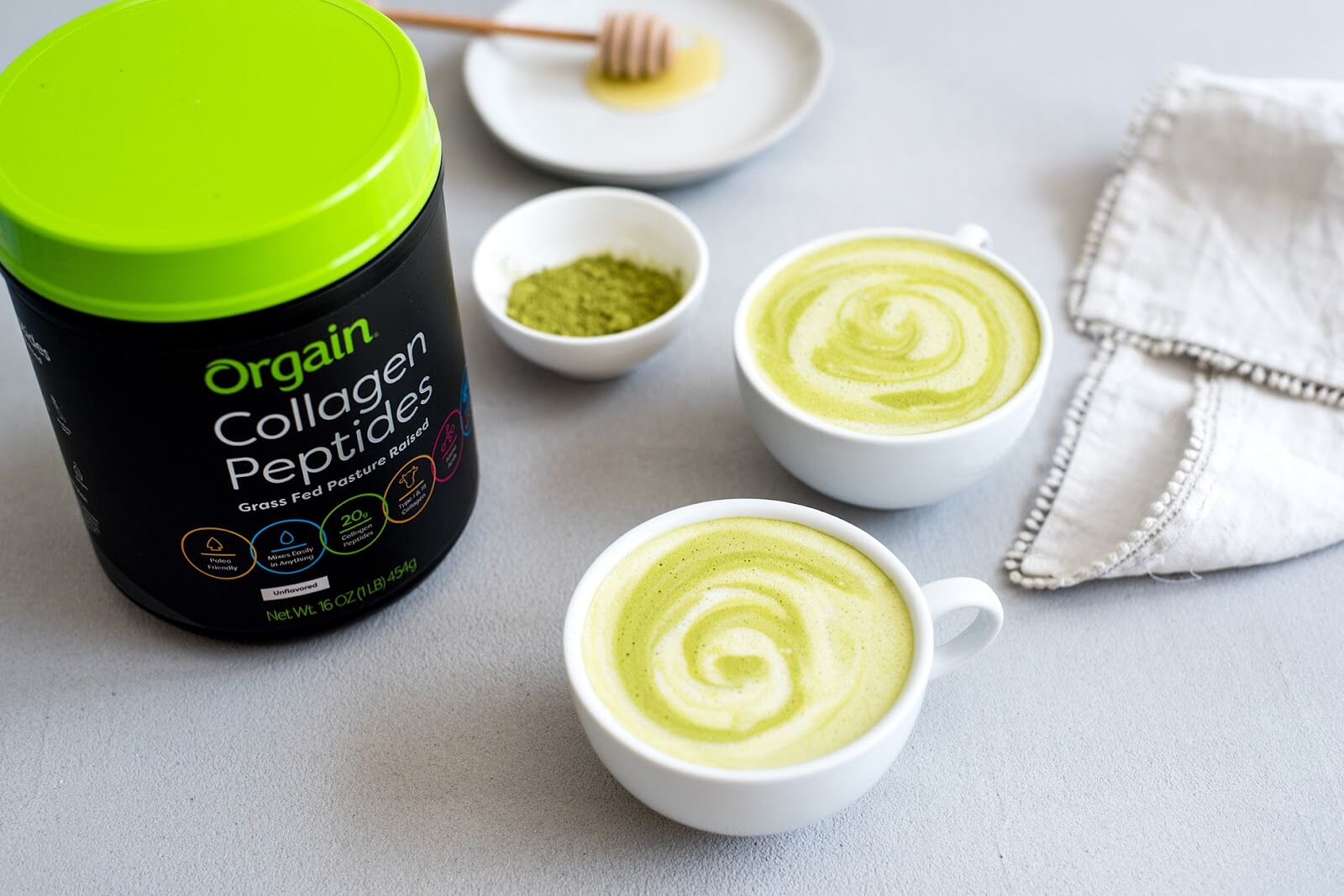 Two cups of matcha lattes with swirled foam sit on a light gray surface next to a folded white cloth. A tub of Orgain Collagen Peptides powder, a small bowl with matcha powder, and a plate with a honey dipper are also visible.