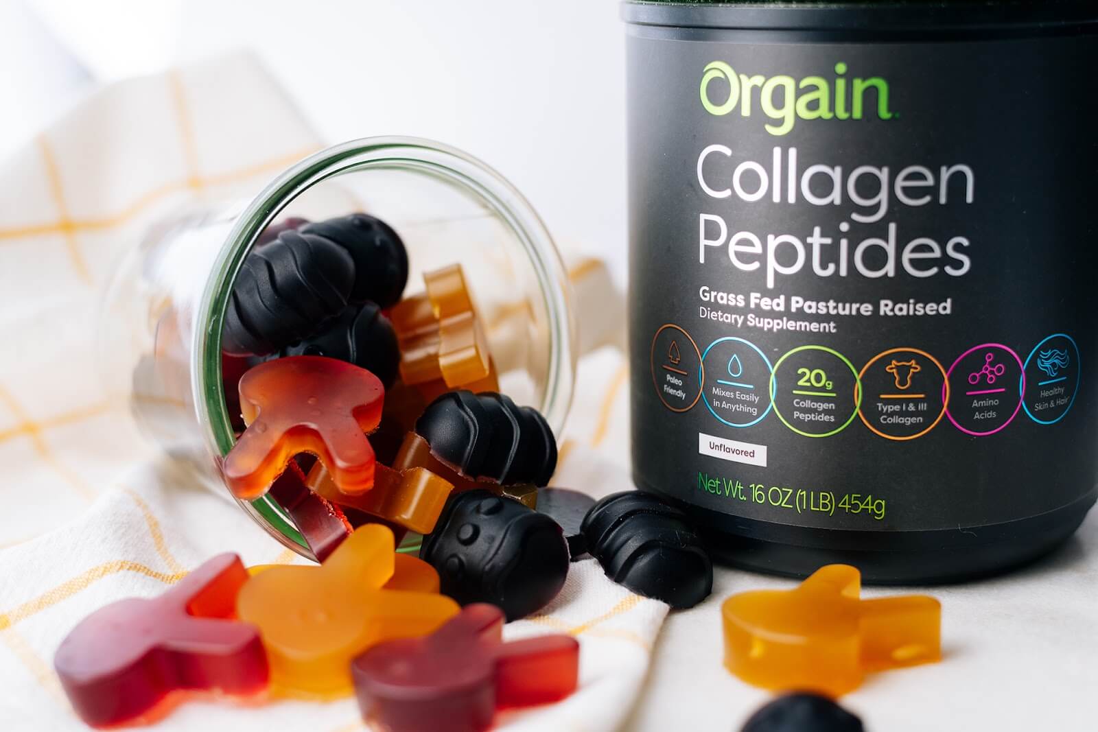 A tipped-over glass jar spills multicolored gummy candies onto a white cloth with yellow stripes. Next to it is a black container of Orgain Collagen Peptides powder.