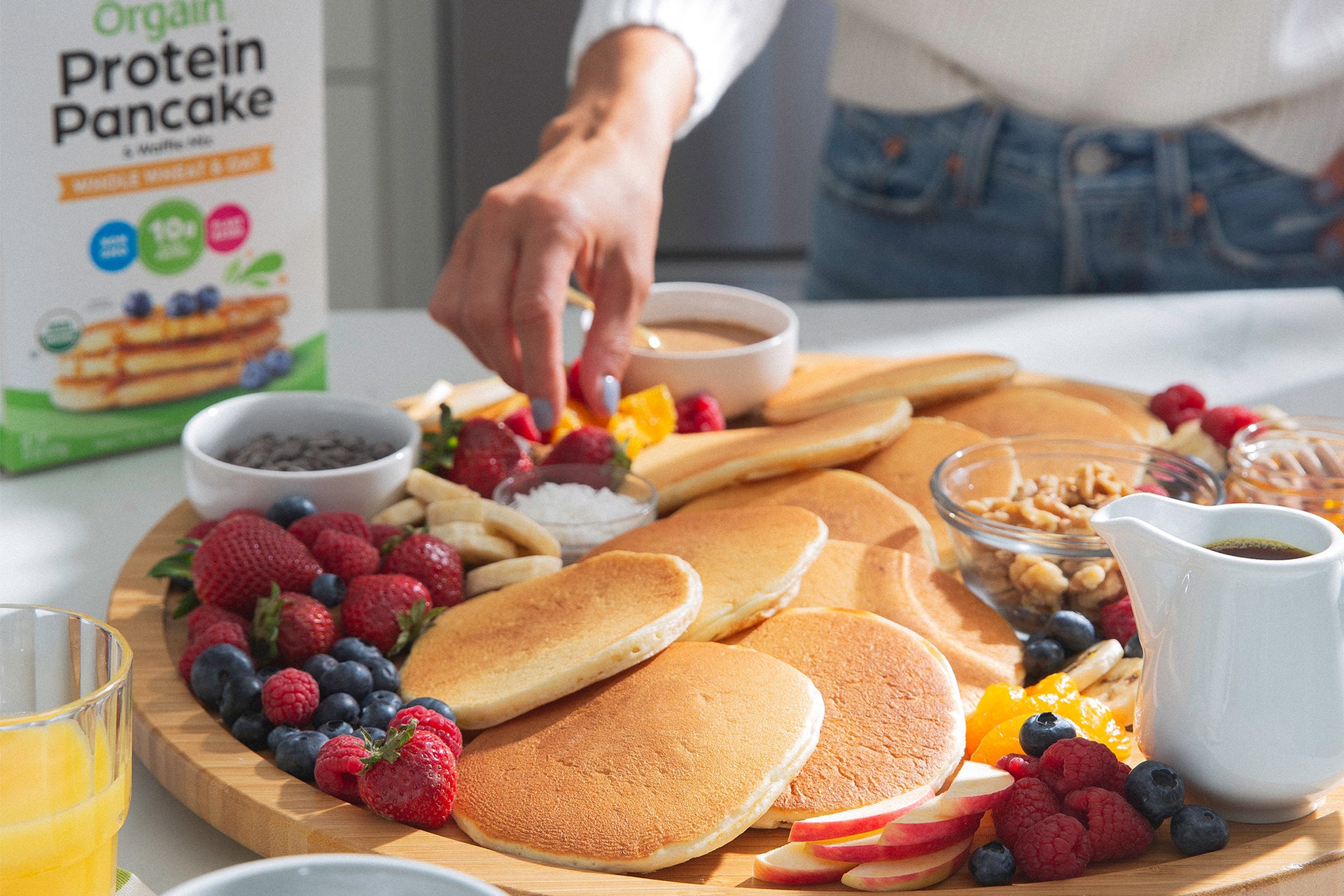 A person arranges items on a large wooden platter filled with pancakes, fresh fruits like berries and orange slices, small bowls of nuts, and syrup. A box of Orgain Protein Pancake mix is visible in the background on a white countertop.