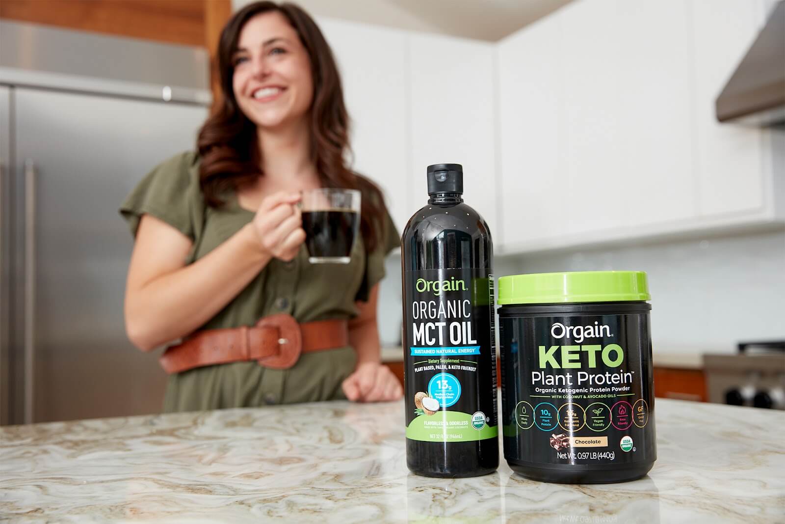 A woman holding a coffee mug stands in a kitchen. In the foreground, Orgain organic MCT oil and Orgain keto plant protein powder are placed on the countertop. The woman, smiling, is out of focus while the products are in sharp focus.