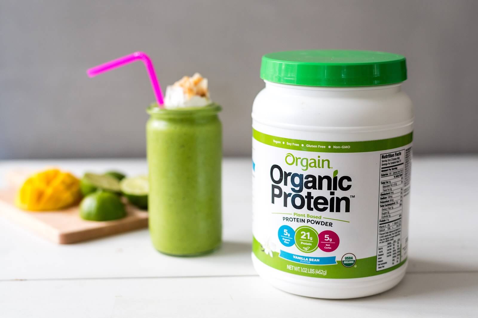 A container of Orgain Organic Protein Powder with a green lid is placed on a white surface. Behind it, there is a green protein shake in a mason jar, topped with whipped cream and a pink straw. Nearby, a cutting board holds a sliced mango and lime.