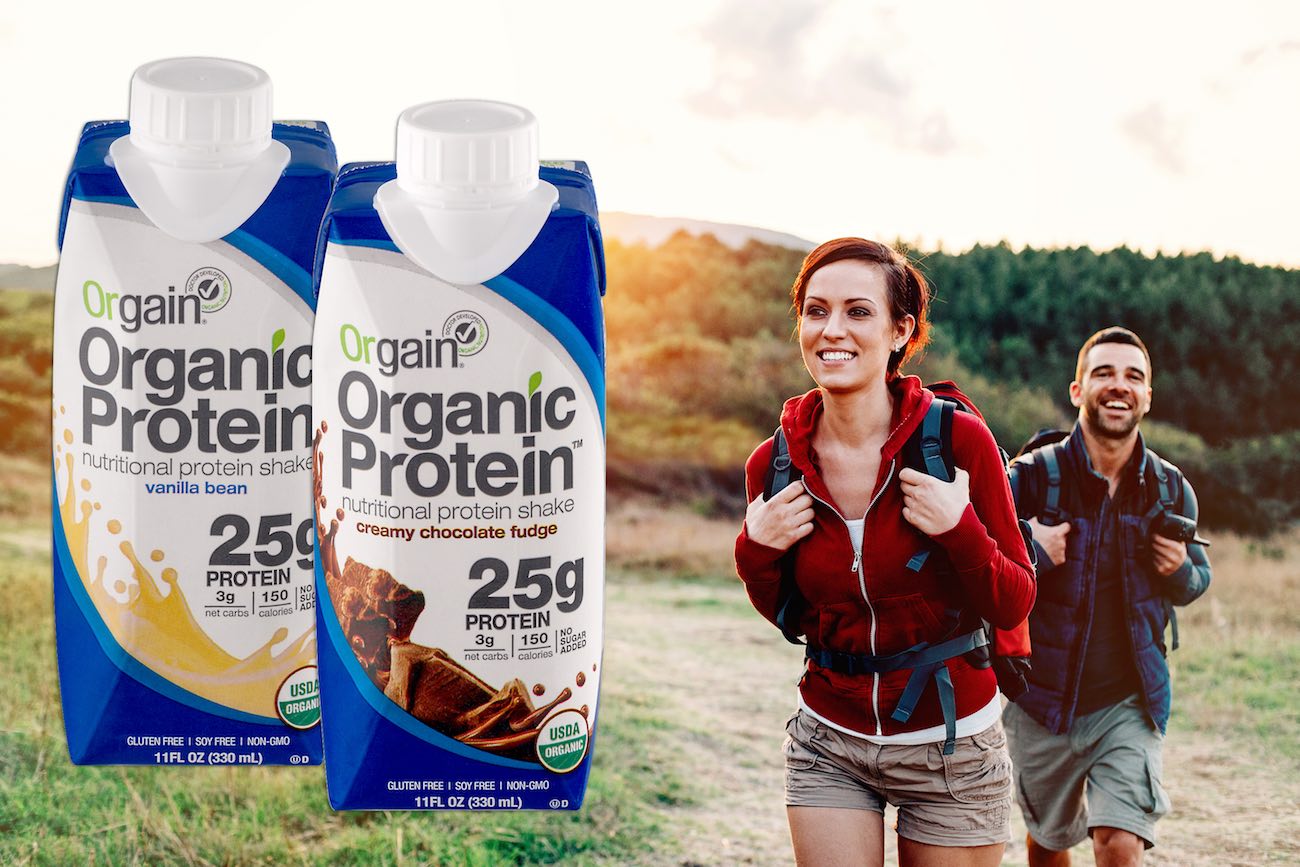 Two people are smiling and hiking outdoors, wearing backpacks. In the foreground, there are two cartons of Orgain Organic Protein nutritional shakes, one vanilla bean flavor and one creamy chocolate fudge flavor, each with 25 grams of protein.