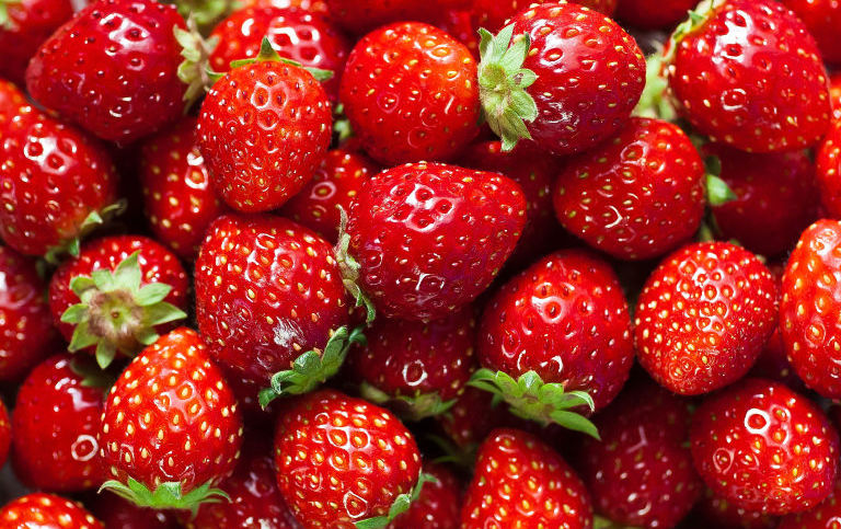A close-up view of a pile of ripe, red strawberries with bright green leaves and small seeds visible on the surface. The strawberries are fresh and glossy, indicating their juiciness and ripeness.