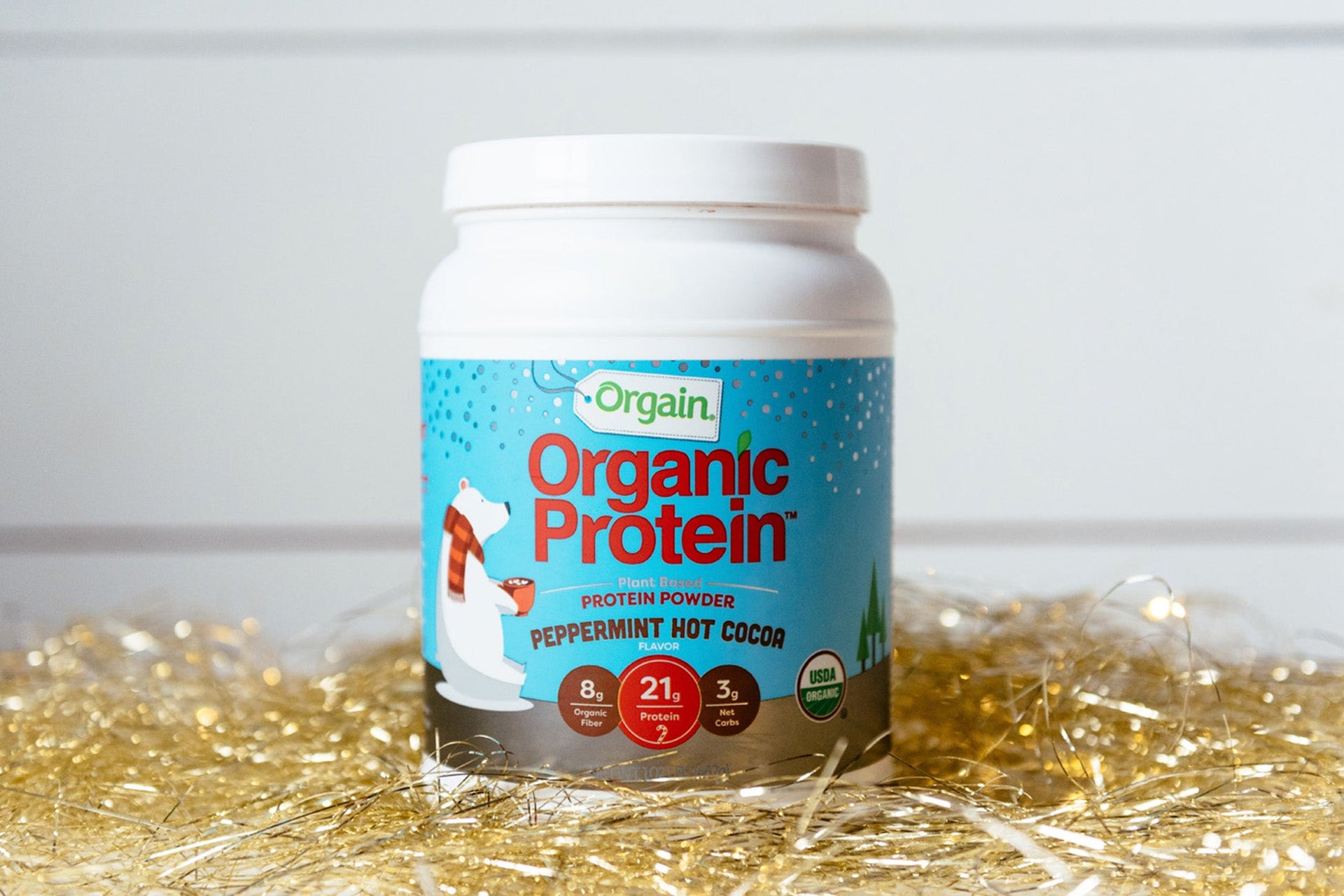 Orgain Organic Protein Powder in Peppermint Hot Cocoa flavor, with a polar bear on the label, among festive decorations.