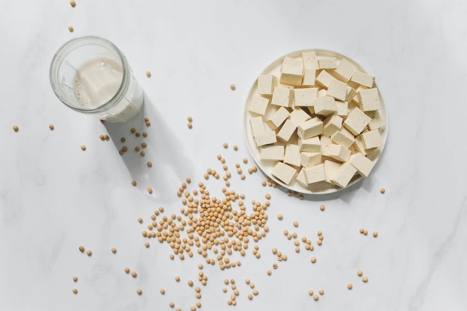 A glass of soy milk, a plate of cubed tofu, and a few scattered soybeans are arranged on a white marble surface. The objects are positioned to the left and center of the frame, with the scattered soybeans creating a pattern.