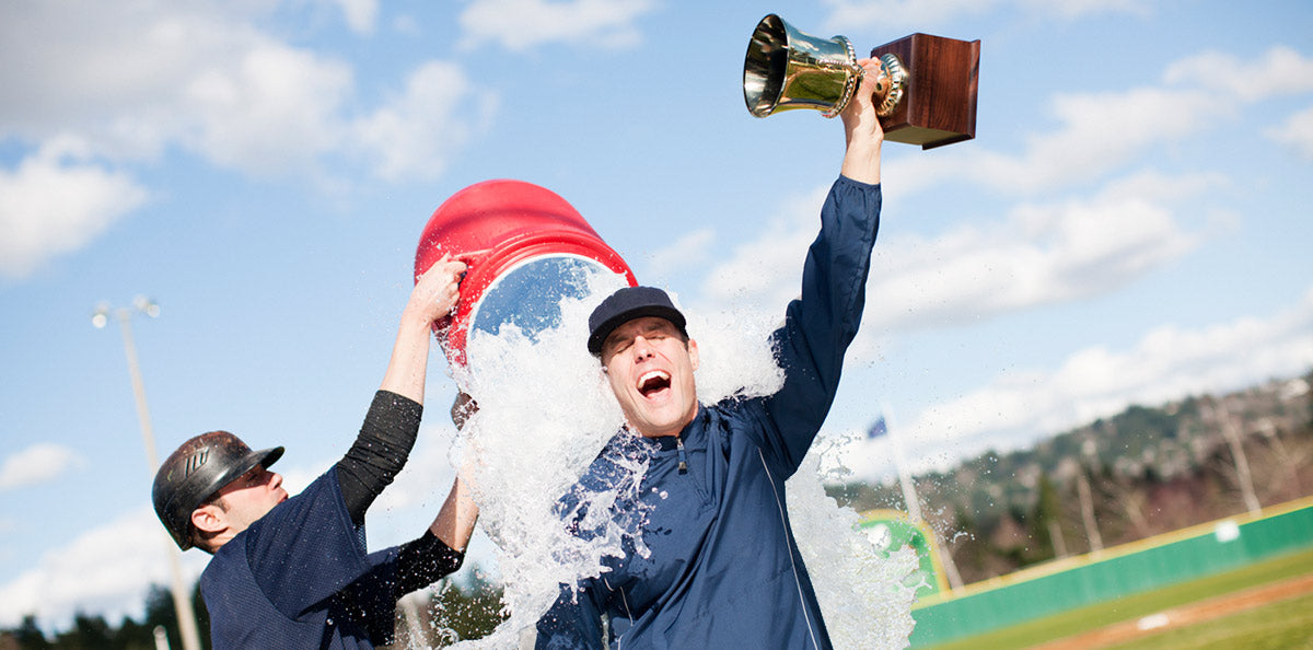 Two baseball players celebrate a victory. One player holds a trophy and raises it triumphantly while being doused with water from a red cooler by the other player. They are on a baseball field with a blue sky and clouds in the background.