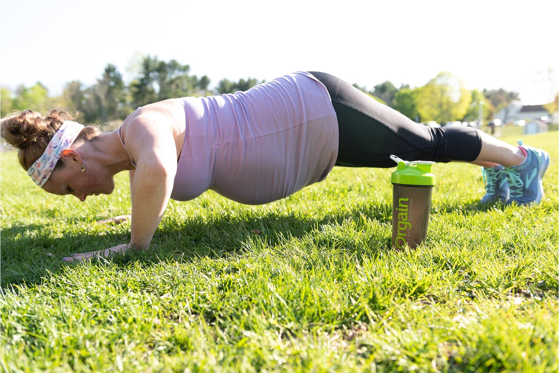 A pregnant woman in athletic wear performs a plank exercise on a grassy field. She wears a headband and bright shoes, and a protein shaker bottle stands next to her. The background features blurred trees and a bright, sunny sky.