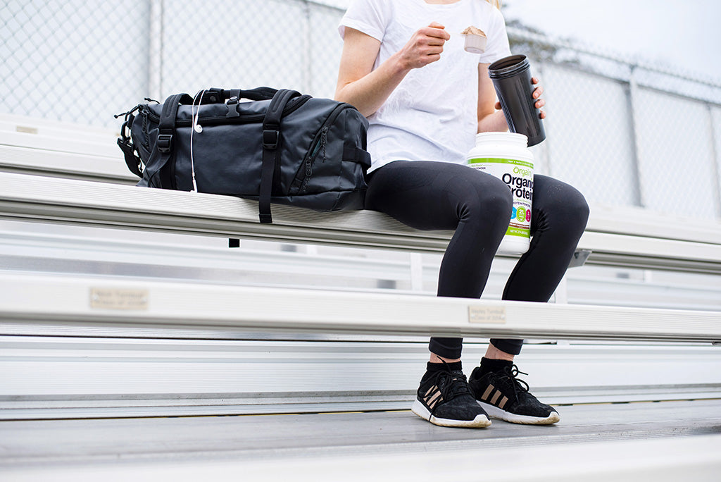 A person wearing a white shirt and black leggings is seated on bleachers. They are holding a black shaker cup and have a container of Orgain Organic Protein powder beside them. A black gym bag is placed on the bench next to the person.