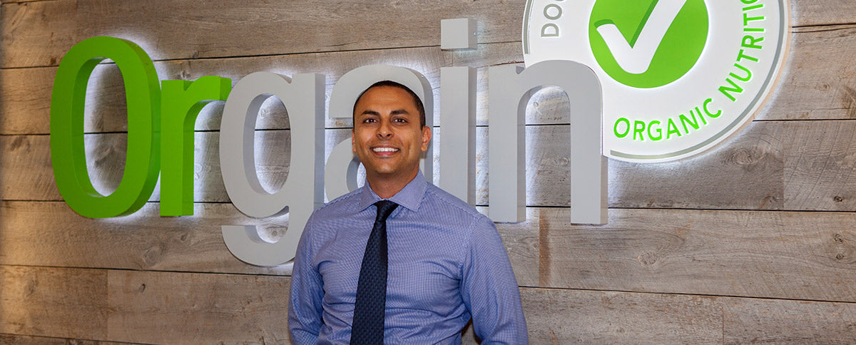 A smiling man in a blue dress shirt and dark tie stands in front of a wooden wall with a large "Orgain" sign and an "Organic Nutrition" logo featuring a green checkmark. The background is well-lit and the man appears to be in a professional setting.