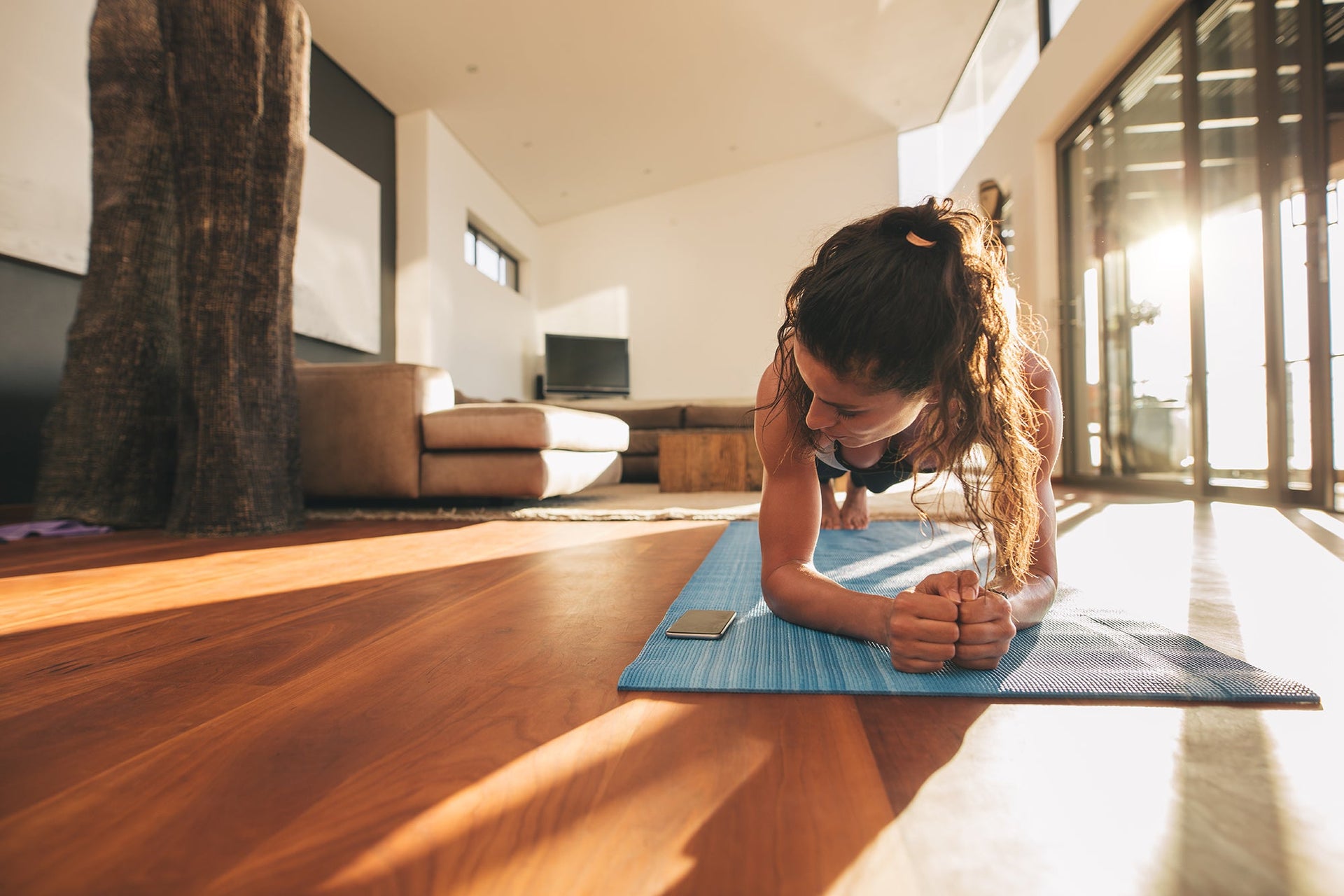 A person performs a plank exercise on a blue yoga mat in a spacious, sunlit living room. The room has large windows, wooden flooring, and modern furniture. The person is focused, with forearms on the mat and legs extended. A phone is placed nearby.