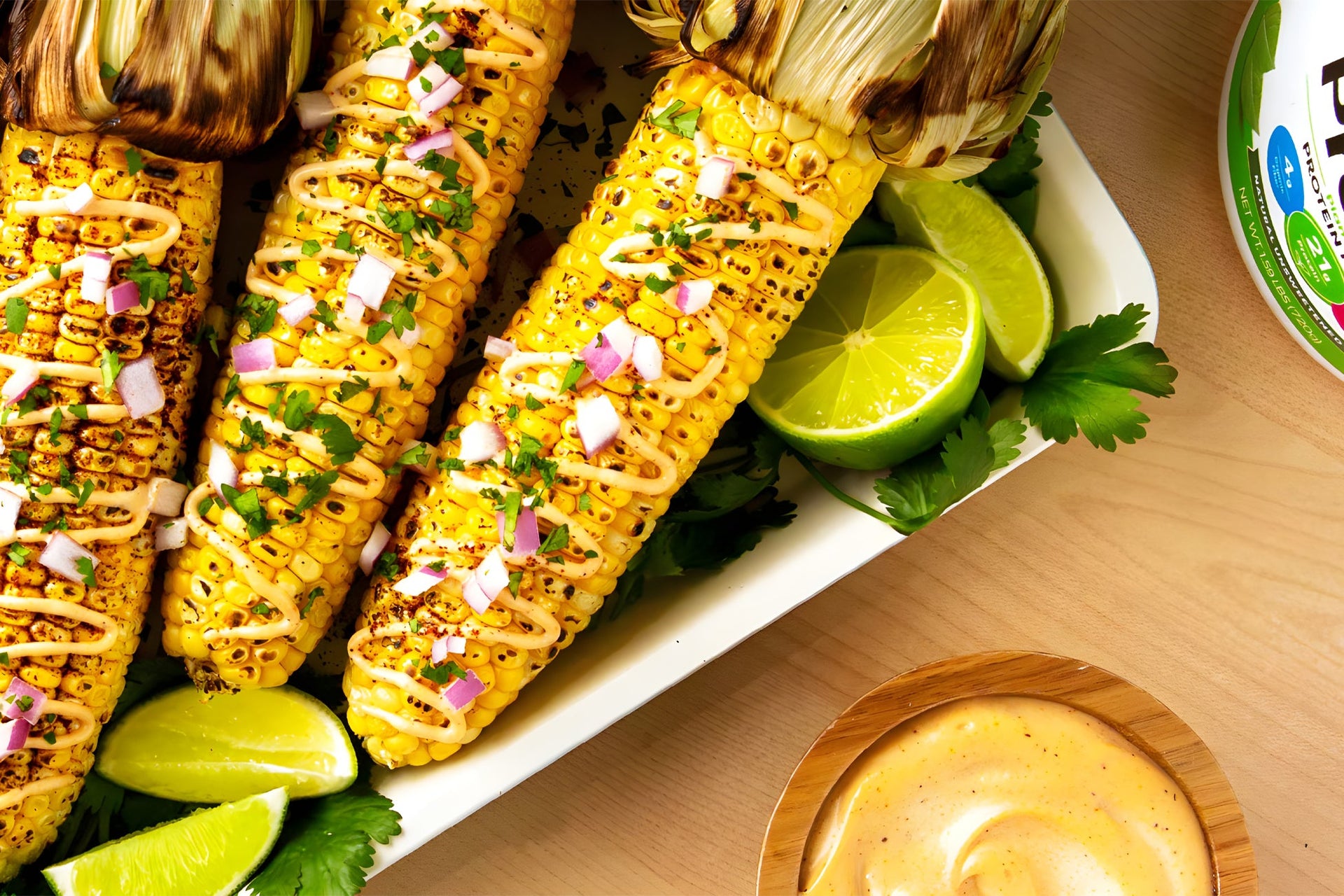 A rectangular white dish holds three grilled corn cobs with creamy sauce, chopped red onions, cilantro, and lime wedges. A bowl of dipping sauce and a partially visible container are nearby.