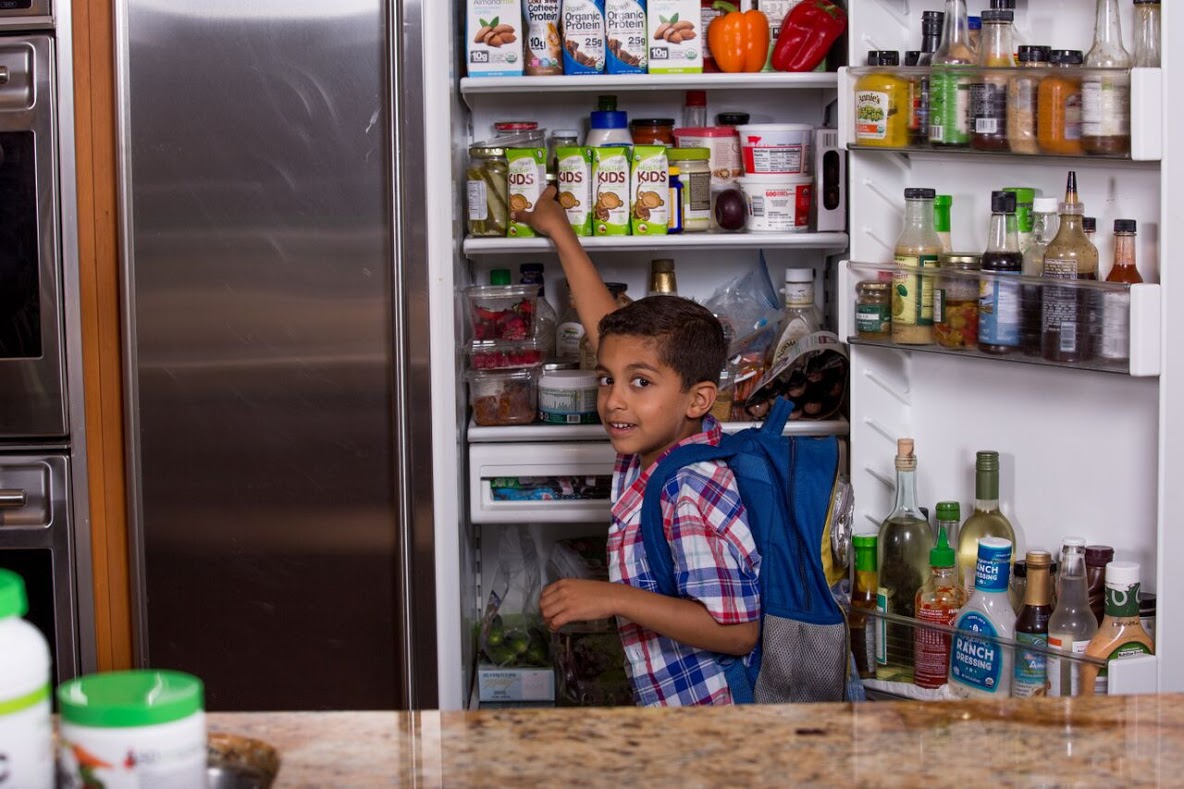 A young boy with a backpack opens a fridge and reaches for a juice box on the top shelf. The refrigerator door is filled with various condiments, sauces, and bottles, while the interior has a variety of food items, including vegetables and juices.