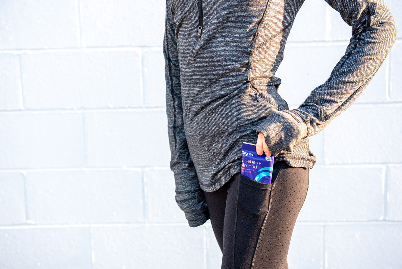 woman standing next to a wall with an orgain protein bar in her pocket