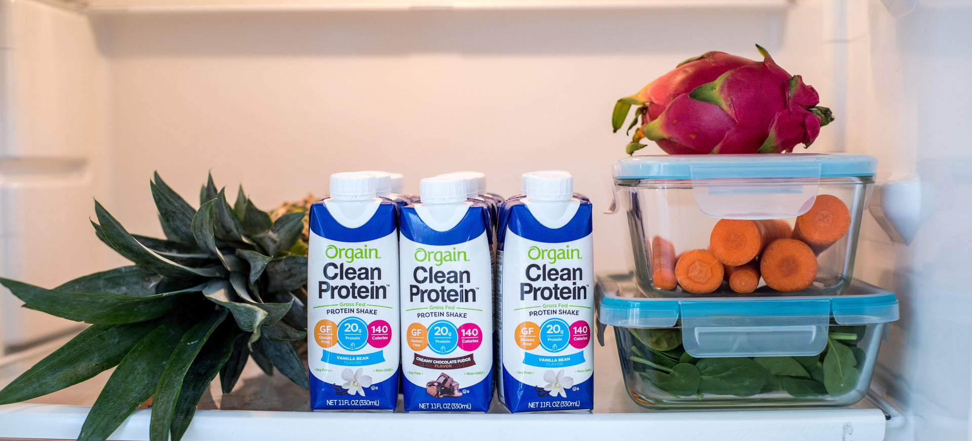 An open refrigerator with three Orgain Clean Protein shake cartons, a whole pineapple, a whole dragon fruit, and two glass containers. One container holds fresh carrots, and the other contains leafy greens.