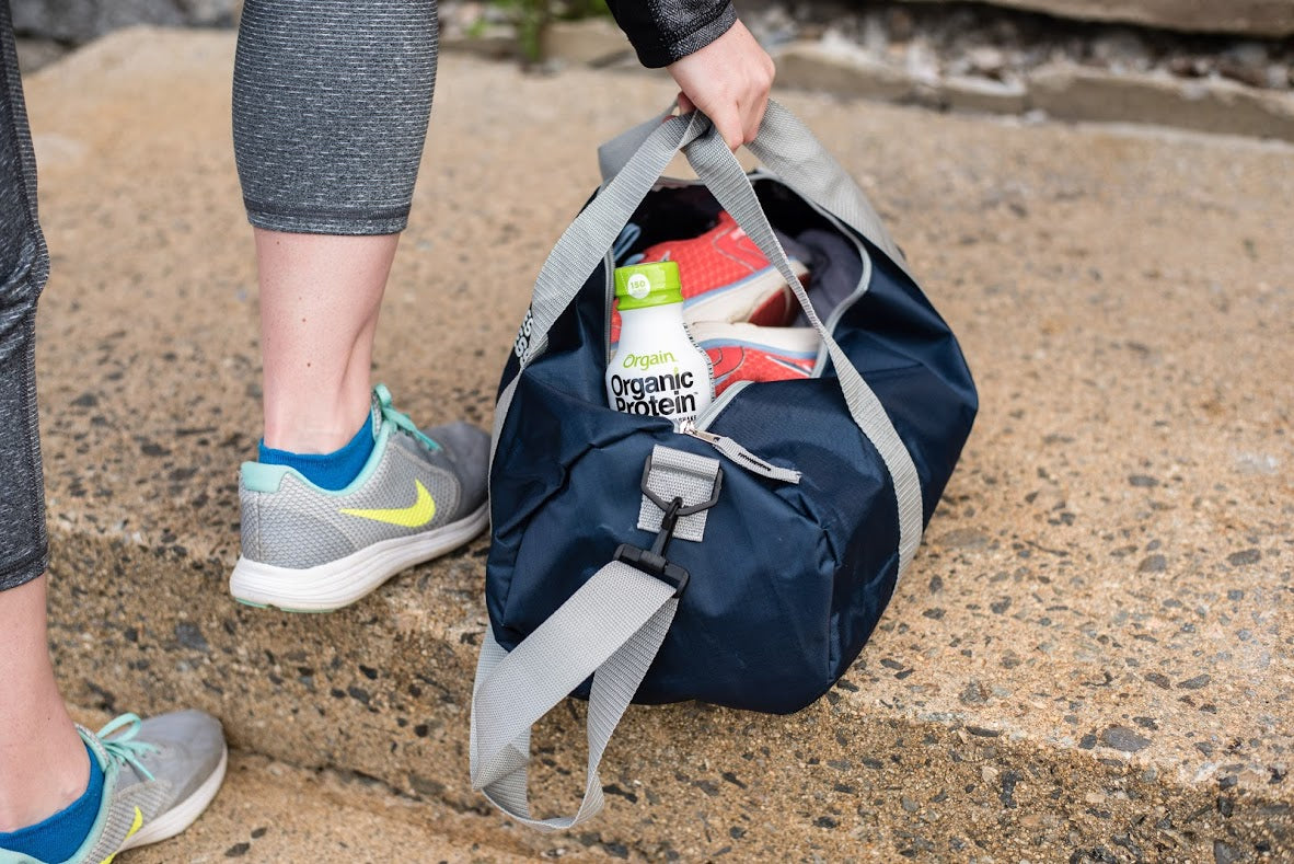 Person wearing gray leggings and teal running shoes placing a navy blue duffel bag on the ground, which contains a pair of red athletic shoes and a carton of organic protein drink. The image appears to depict a pre or post-workout setting.